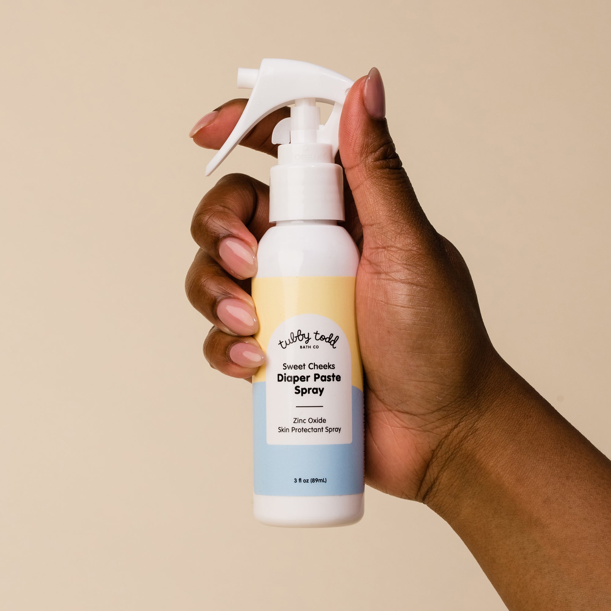 Hand holding the Diaper Paste Spray on the cream color background.